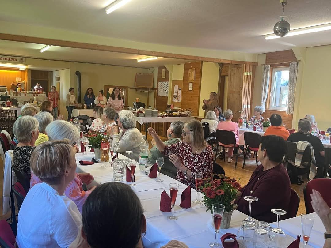 Eine Gruppe von Menschen sitzt an Tischen in einem Esszimmer, einige stehen und unterhalten sich. Blumensträuße, Weingläser und Flaschen schmücken die Tische. Eine Frau in einem Blumenkleid gestikuliert, während andere in Gespräche vertieft sind.