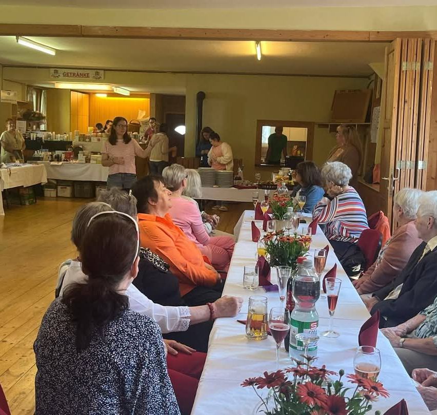 Eine Gruppe älterer Frauen sitzt an einem langen Tisch, einige mit Getränken, Blumen und Tellern. Eine Frau steht vorne, andere bereiten Essen vor. Ein Schild heißt Getranke.