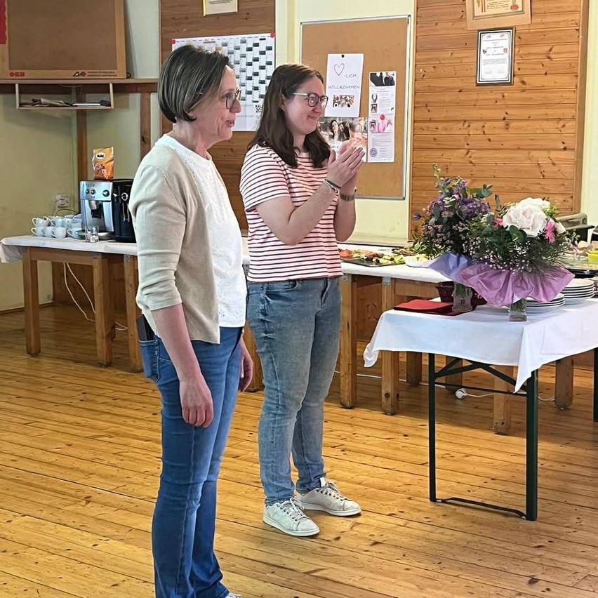 Zwei Frauen stehen in einem Raum, eine klatscht, die andere lächelt. Hinter ihnen steht ein Tisch mit Essen und Blumen. In der Nähe befinden sich eine Kaffeemaschine und ein Bulletin Board.