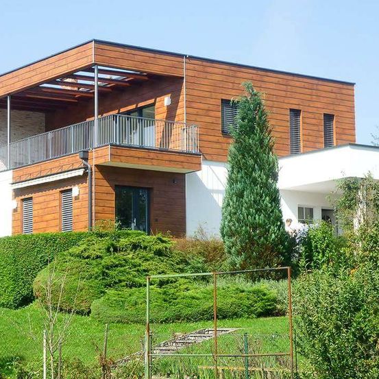 Ein modernes Haus mit Holzfassade und Balkon, umgeben von einem gepflegten Garten mit verschiedenen Sträuchern und Bäumen.