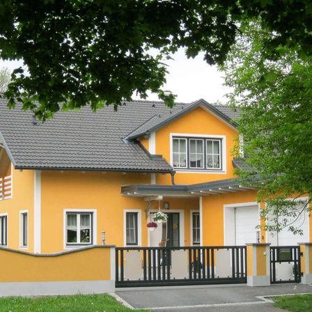 Ein hellgelbes Haus mit grauem Dach und weißen Fenstern, umgeben von viel Grün. Vorne befindet sich ein schwarzes Tor, eine weiße Wand und ein hängender Blumentopf.