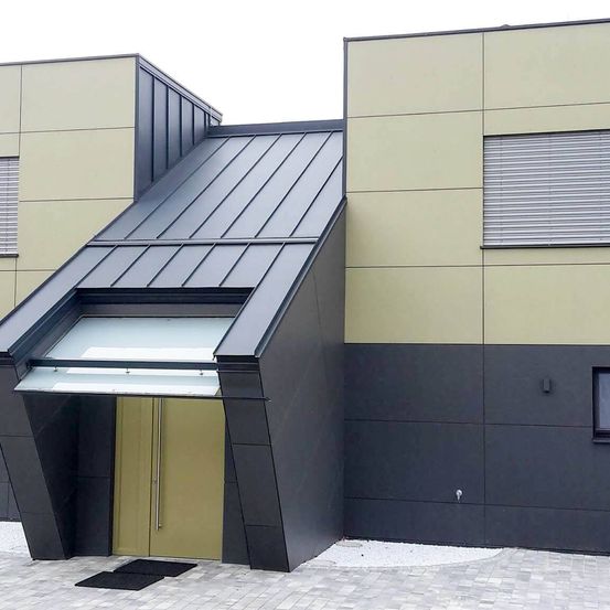 Ein modernes Gebäude mit schwarz-beige Außenfassade, das eine verglaste Überdachung über dem Eingang hat.