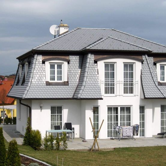 Ein modernes weißes Haus mit einem grauen Dach und mehreren Fenstern. Eine Satellitenschüssel befindet sich auf dem Dach. Das Haus hat eine Terrasse mit Tisch und Stühlen.