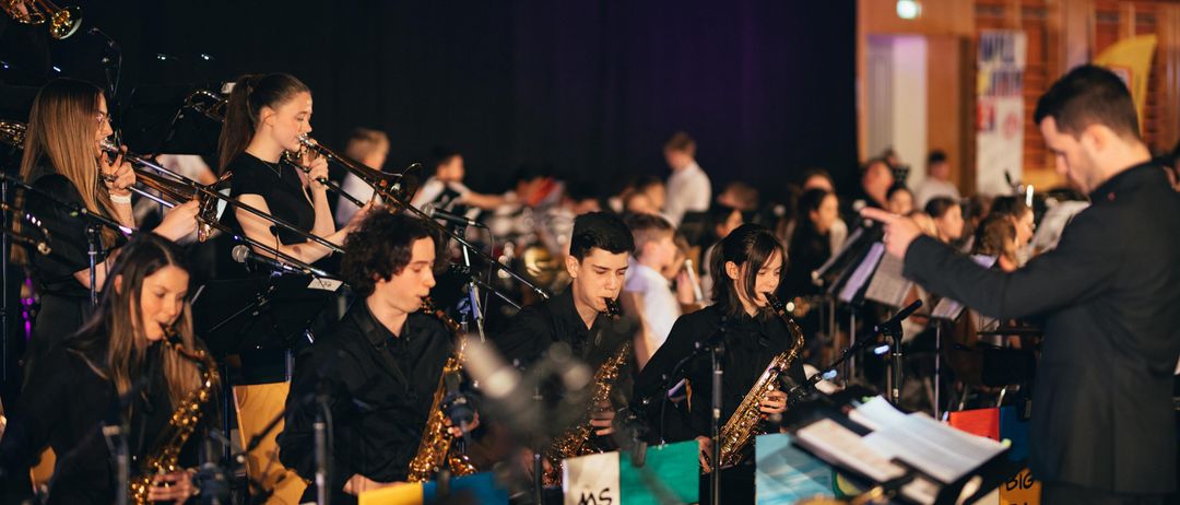 Eine Gruppe von Musikern spielt auf der Bühne mit verschiedenen Instrumenten, darunter Saxophone und Posaunen. Das Publikum sitzt vor ihnen.