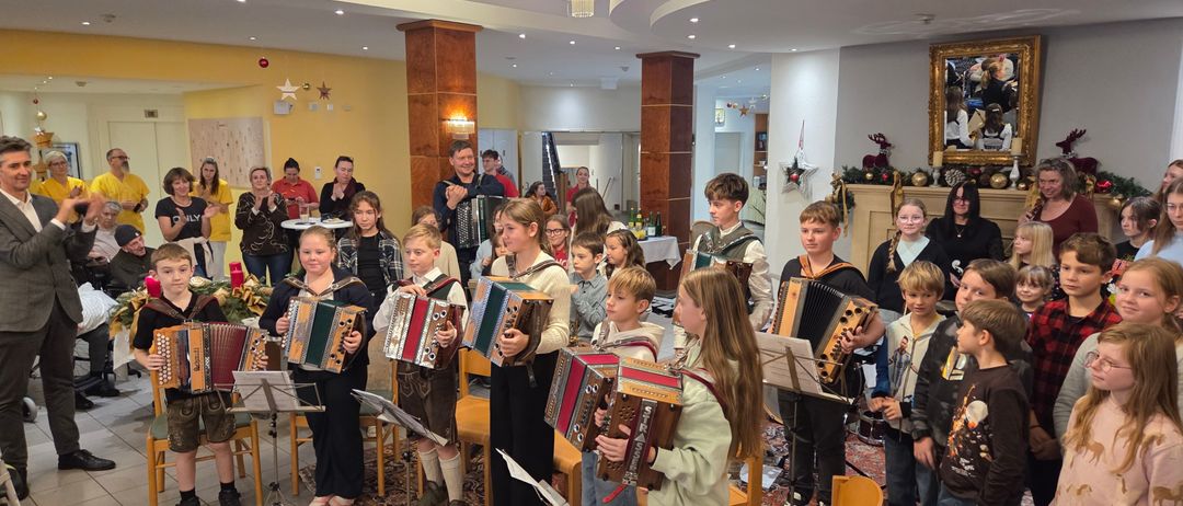 Eine Gruppe von Kindern spielt Akkordeons in einem dekorierten Raum. Einige tragen traditionelle Kleidung, und andere stehen hinter ihnen und klatschen. Eine Kristallkronleuchter und Weihnachtsdekorationen schmücken den Hintergrund.