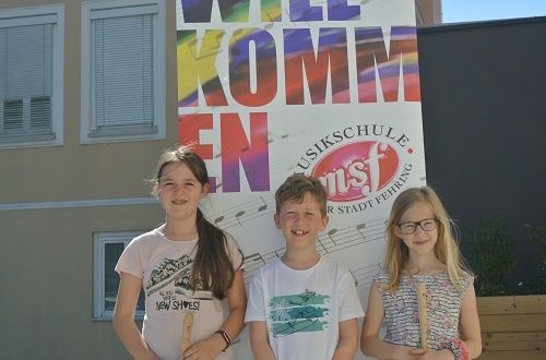 Drei Kinder stehen zusammen mit Musikinstrumenten vor einem Schild der Musikschule.