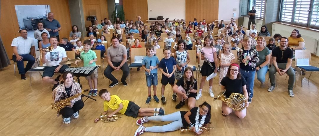 Eine große Gruppe von Kindern und Erwachsenen in einem Saal, einige spielen Instrumente wie Flöten und Trompeten. Ein Mann sitzt lächelnd vorne.