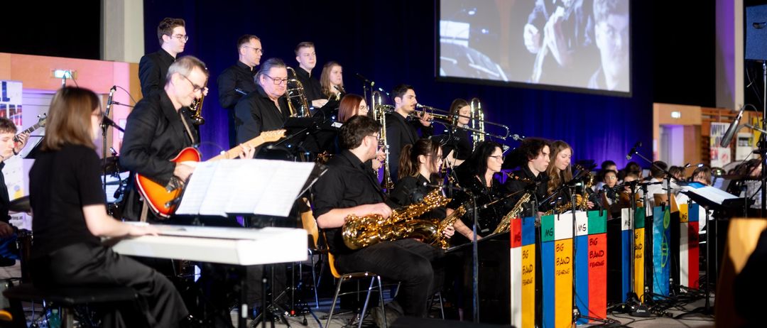 Eine Jazzband spielt auf der Bühne mit einer großen Leinwand im Hintergrund, die einen Gitarristen zeigt. Die Band besteht aus Blech-, Holzblas- und Tasteninstrumenten.