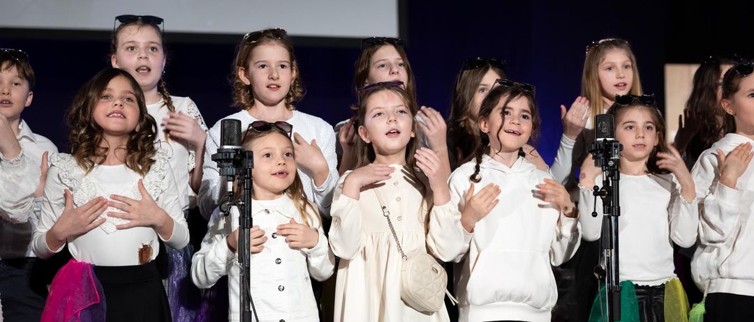 Eine Gruppe junger Mädchen, in verschiedenen Outfits gekleidet, steht auf der Bühne vor Mikrofonen und klatscht in die Hände. Sie scheinen bei einer Veranstaltung aufzutreten, mit einer Leinwand im Hintergrund.