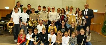 Eine Gruppe von Kindern und Erwachsenen posiert für ein Foto in einem Raum mit Holzböden. Sie halten Musikinstrumente. Ein Klavier und eine Topfpflanze befinden sich auf der linken Seite.