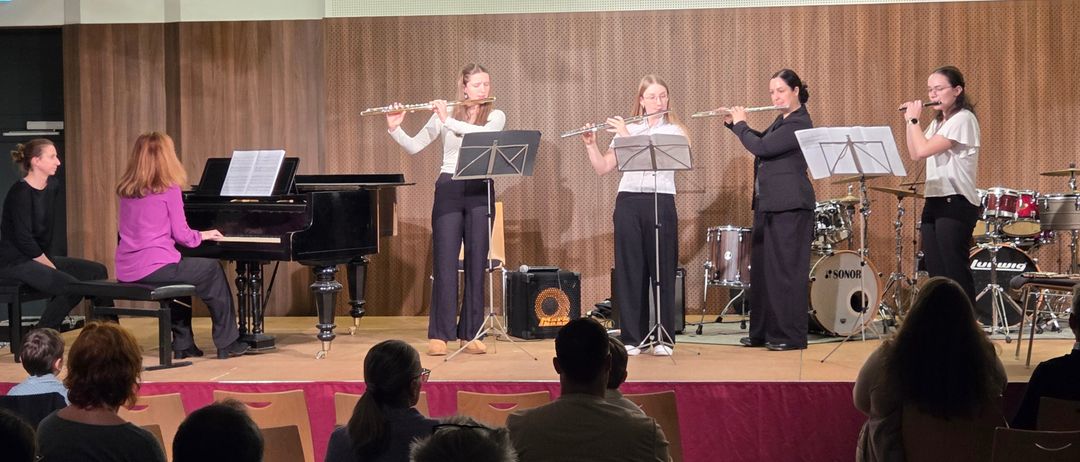 Drei Frauen spielen Flöten auf einer Bühne mit Klavier und Schlagzeug im Hintergrund. Das Publikum sitzt vor ihnen und beobachtet die Aufführung.