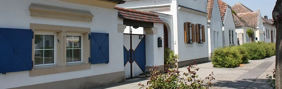 Ein weißes Gebäude mit blauen Fensterläden und einer blauen Tür, begleitet von grünen Büschen und einem Betonpfad.