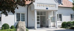 Ein weißes Gebäude mit roten Dachziegeln. Es hat den Text 'GEMEINDEAM' oben und die Nummer '7' darunter. Der Eingang ist offen mit einer Tür. Vorne steht eine Bank.