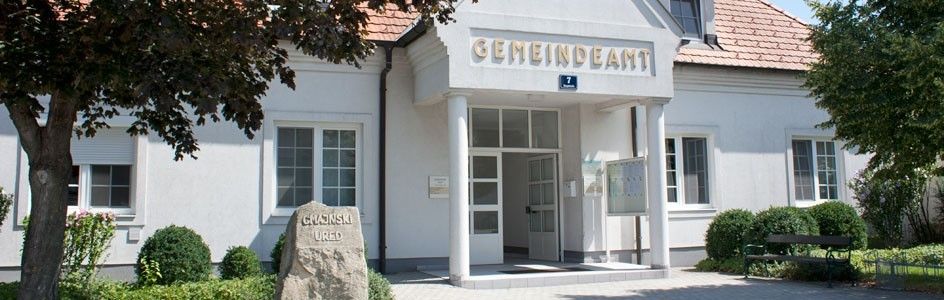 Ein weißes Gebäude mit roten Dachziegeln. Es hat den Text 'GEMEINDEAM' oben und die Nummer '7' darunter. Der Eingang ist offen mit einer Tür. Vorne steht eine Bank.