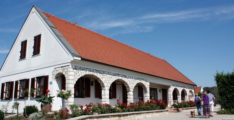 Ein weißes Haus mit rotem Dach und Bogenfenstern steht vor einem blauen Himmel. Zwei Frauen spazieren mit einem Hund vor dem Haus.