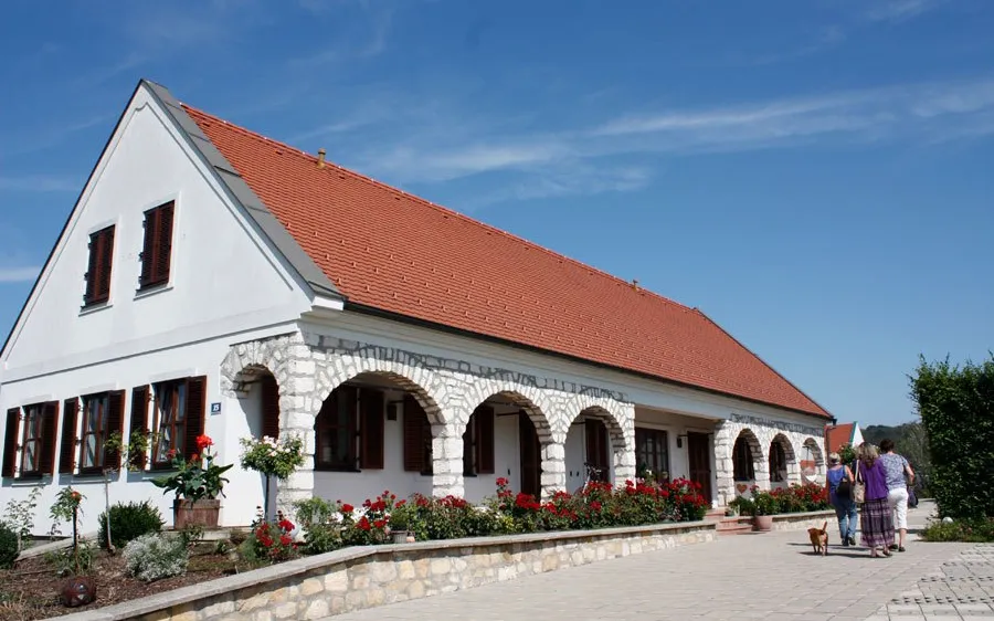 Ein weißes Haus mit rotem Dach und Bogenfenstern steht vor einem blauen Himmel. Zwei Frauen spazieren mit einem Hund vor dem Haus.