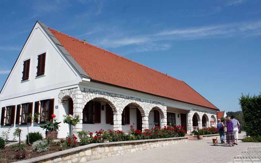 Ein weißes Haus mit rotem Dach und Bogenfenstern steht vor einem blauen Himmel. Zwei Frauen spazieren mit einem Hund vor dem Haus.