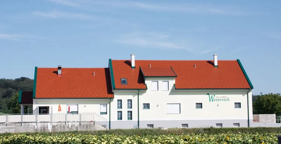 Ein weißes Gebäude mit rotem Dach und grüner Einfassung steht inmitten eines Sonnenblumenfeldes unter einem klaren blauen Himmel.