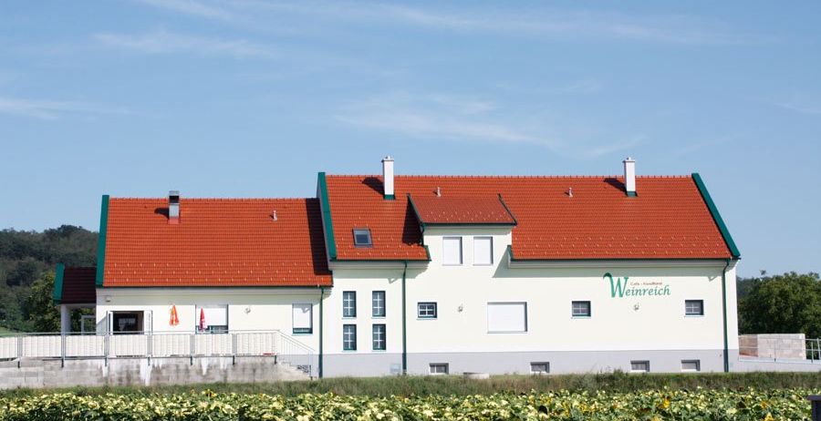 Ein weißes Gebäude mit rotem Dach und grüner Einfassung steht inmitten eines Sonnenblumenfeldes unter einem klaren blauen Himmel.