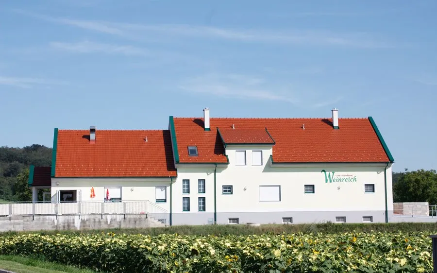 Ein weißes Gebäude mit rotem Dach und grüner Einfassung steht inmitten eines Sonnenblumenfeldes unter einem klaren blauen Himmel.