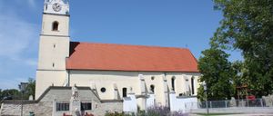 Eine traditionelle Kirche mit rotem Dach und einem Turm mit einer Uhr, umgeben von Grün und Blumen. Eine Statue ziert die Vorderseite.