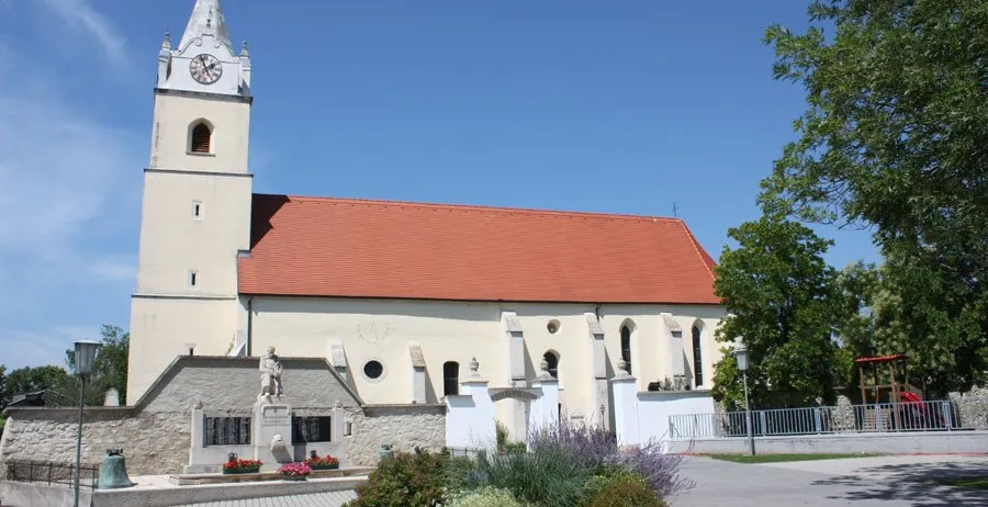 Eine traditionelle Kirche mit rotem Dach und einem Turm mit einer Uhr, umgeben von Grün und Blumen. Eine Statue ziert die Vorderseite.