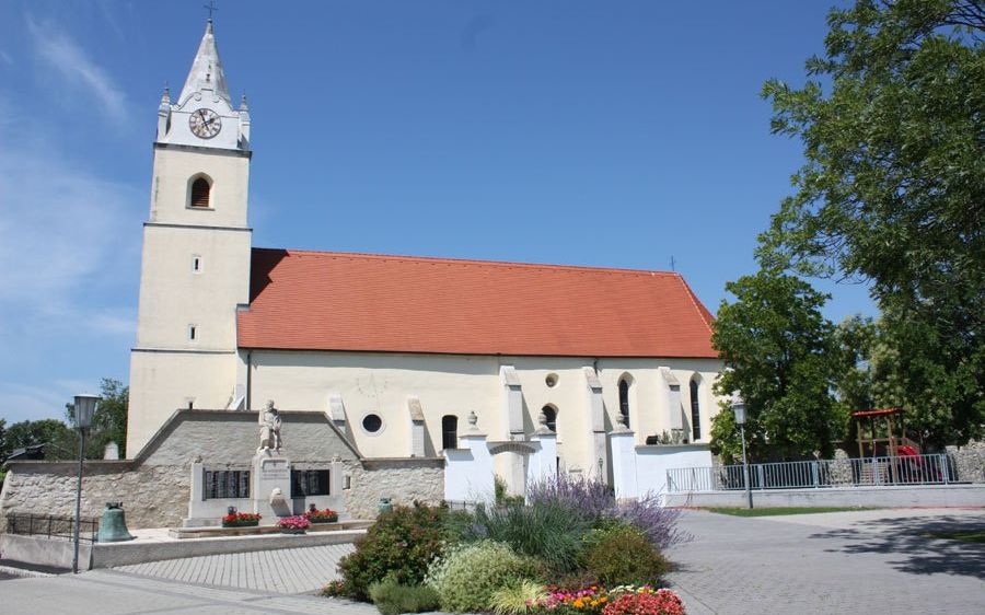 Eine traditionelle Kirche mit rotem Dach und einem Turm mit einer Uhr, umgeben von Grün und Blumen. Eine Statue ziert die Vorderseite.