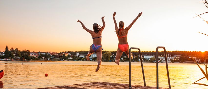 Zwei Frauen in Badeanzügen springen bei Sonnenuntergang von einem Steg in einen See, Arme erhoben. Hinter ihnen liegt eine ruhige Stadtlandschaft mit Häusern und Bäumen.