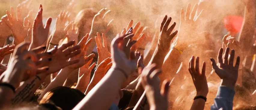 Eine Menschenmenge, die die Hände hebt, wahrscheinlich bei einem Konzert oder einer Veranstaltung. Die Hände sind verschwommen, und einige tragen Armbänder. Die Menschen stehen eng beieinander.