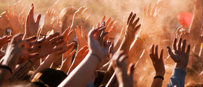 Eine Menschenmenge, die die Hände hebt, wahrscheinlich bei einem Konzert oder einer Veranstaltung. Die Hände sind verschwommen, und einige tragen Armbänder. Die Menschen stehen eng beieinander.
