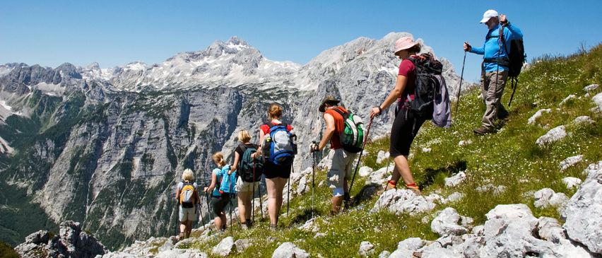 Eine Gruppe von Wanderern mit Rucksäcken steigt einen grasigen Hang mit felsigem Gelände in den Bergen hinauf. Sie benutzen Trekkingstöcke und tragen Mützen.