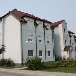 Ein modernes Mehrfamilienhaus mit roten Dächern und weißen Wänden steht an einem sonnigen Tag, umgeben von Grün und einer Straßenlaterne.