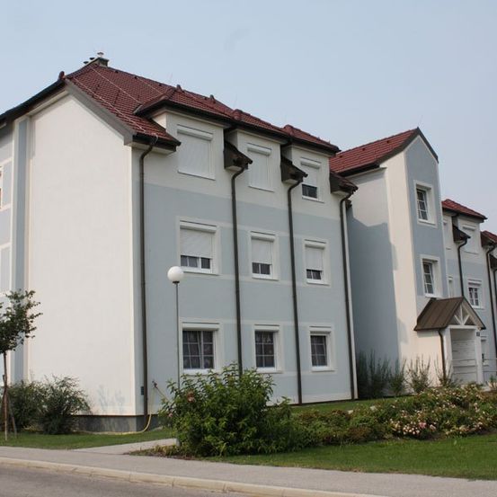 Ein modernes Mehrfamilienhaus mit roten Dächern und weißen Wänden steht an einem sonnigen Tag, umgeben von Grün und einer Straßenlaterne.
