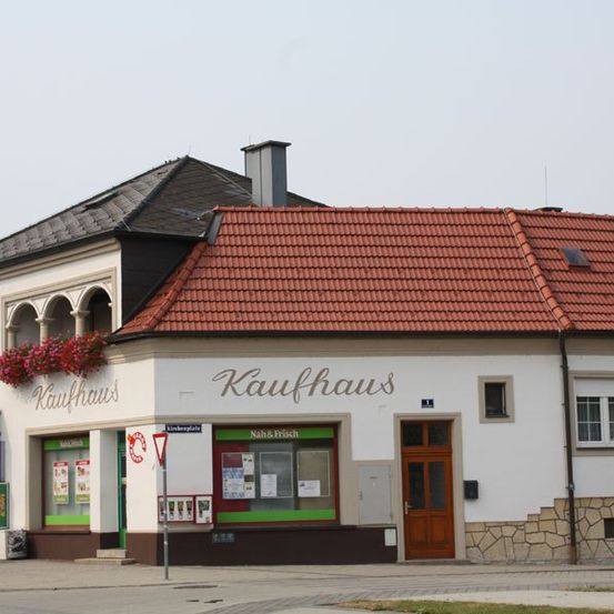 Ein zweistöckiges Gebäude mit rotem Dach hat ein Schild mit der Aufschrift 'Kaufhaus'. Es hat zwei Eingänge mit Schildern darüber. Ein Fahrrad ist links geparkt.