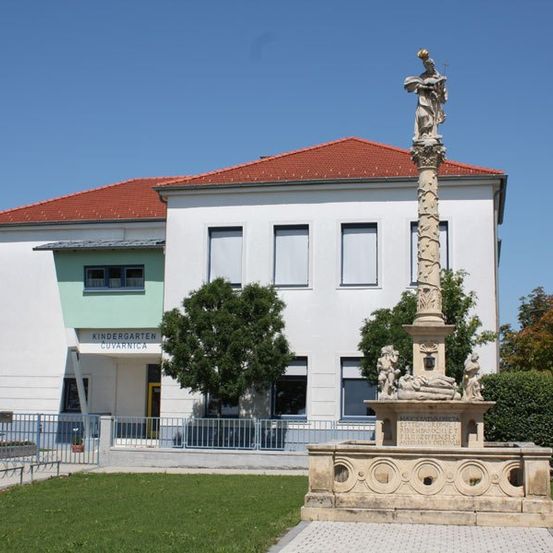 Ein weißes Gebäude mit rotem Dach und einer Tafel mit der Aufschrift 'Kindergarten Cuvanica' steht hinter einem großen Steinmonument. Das Monument zeigt eine Statue und einige Schnitzereien. Bäume und Büsche umgeben das Monument.