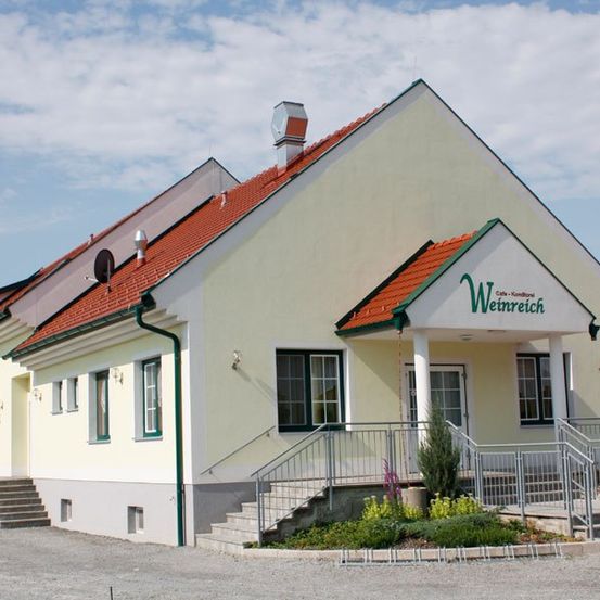 Ein weißes Gebäude mit rotem Dach namens Weinreich. Es hat einen vorderen Eingang mit Treppen und Geländern und einen Garten mit Pflanzen davor.