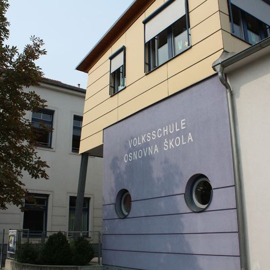 Ein Gebäude mit gelben und lila Wänden trägt die Aufschrift 'Volksschule Osnovna Skole' an seiner Vorderseite. Ein Baum steht davor.