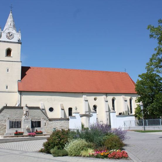Eine weiße Kirche mit rotem Dach und Glockenturm. Eine Statue steht vorne. Vor der Kirche sind Pflanzen und Blumen. Bäume und ein Zaun befinden sich auf der rechten Seite.