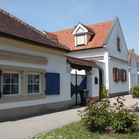 Ein weißes Gebäude mit rotem Dach und blauen Fensterläden. Es hat eine geschlossene Eingangstür und zwei Fenster. Vor dem Eingang steht ein Mülleimer.