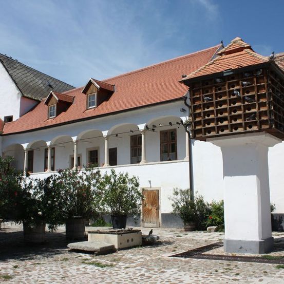 Ein traditionelles europäisches Haus mit Terrakotta-Dächern und Holzelementen. Tauben sitzen auf einem hölzern Taubenschlag. Pflanzen sind um das Haus herum aufgestellt.
