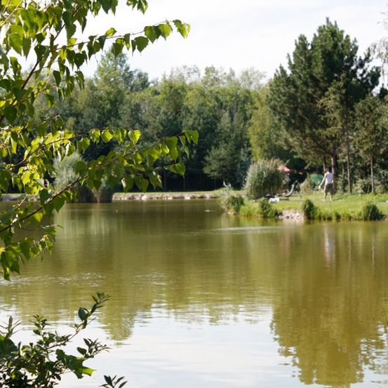 Ein ruhiger See, umgeben von üppigem grünem Laub und Büschen. Zwei Personen stehen in der Nähe des Wassers, und ein roter Schirm ist in der Nähe.