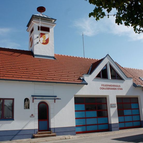 Ein weißes Gebäude mit einem roten Dach und einem Turm darauf, es hat zwei rote Garagentore und blaue Verzierungen, mit einem deutschen Schild an der Vorderseite, das 'Feuerwehrhaus Ognjobranski Stan' besagt.