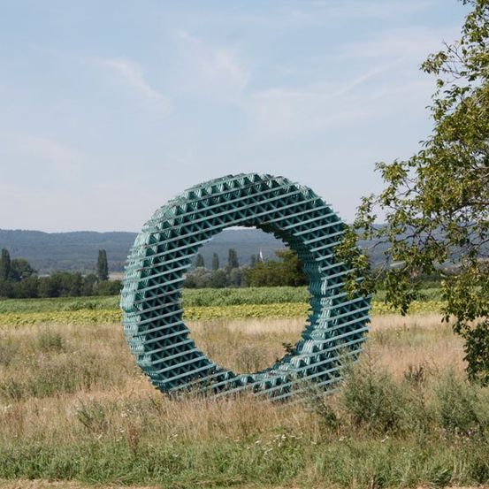 Eine große kreisförmige Struktur aus grünen Kisten steht auf einem weitläufigen, offenen Feld unter einem blauen Himmel mit einigen Wolken.