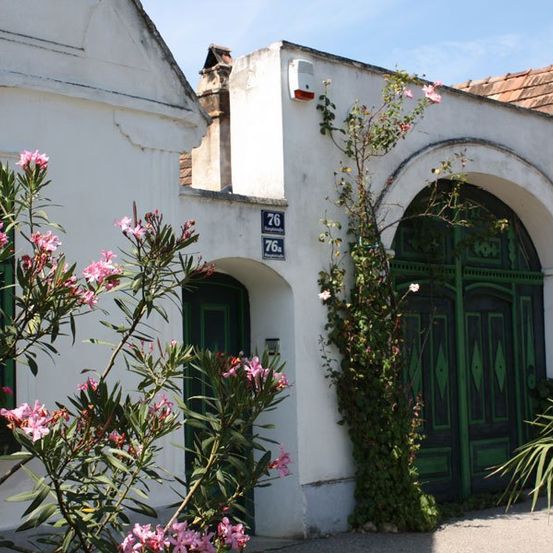Ein weißes Gebäude mit grünen Türen und rosa Blumen davor, Nummer 26 an der Wand und einem roten Dach.