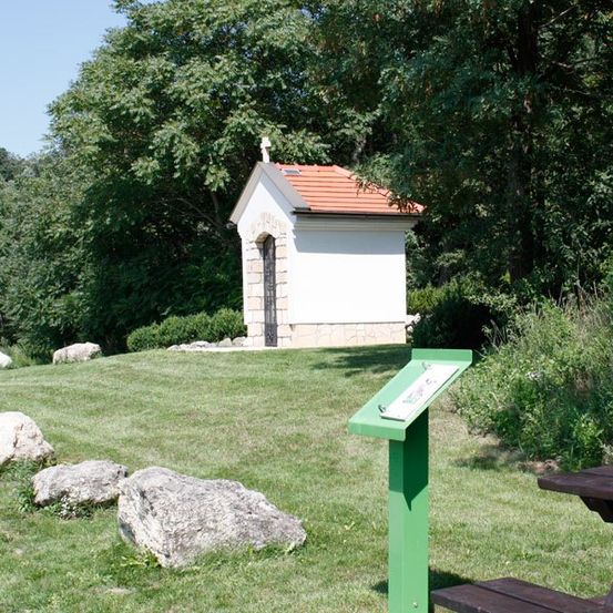 Eine kleine Kapelle mit rotem Dach steht auf einer Rasenfläche, mit umliegenden Steinen und einem grünen Informationspfosten in der Nähe.