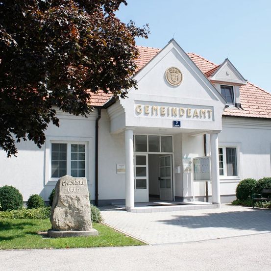 Ein weißes Gebäude mit rotem Dach hat ein Schild mit der Aufschrift Gemeindeamt über dem Eingang. Ein Steinpfeiler mit den Worten Chaonski Ured steht auf dem Rasen.
