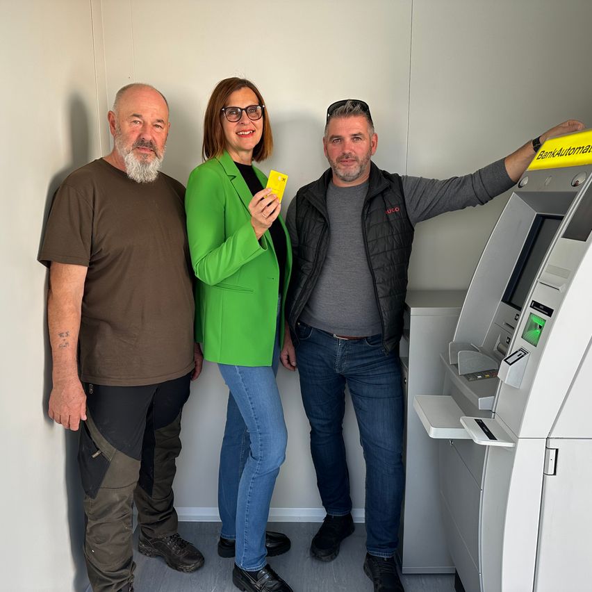Drei Personen stehen gemeinsam vor einem Bankautomaten. Der Mann auf der linken Seite trägt ein braunes T-Shirt. Die Frau in der Mitte hält eine gelbe Karte. Der Mann auf der rechten Seite trägt eine schwarze Weste.