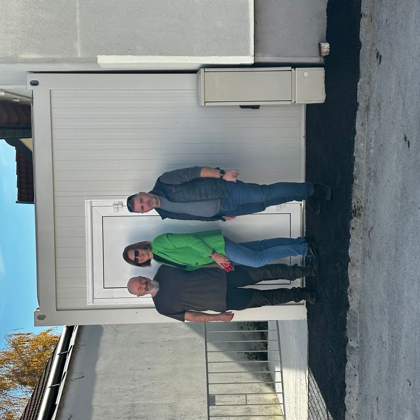 Drei Personen stehen vor einem Haus mit einer Garagentür und lächeln für die Kamera. Ein Mann lehnt sich an die Wand, eine Frau steht mit einem roten Gegenstand in der Hand. Der dritte Mann kniet, und ein Baum ist links sichtbar.