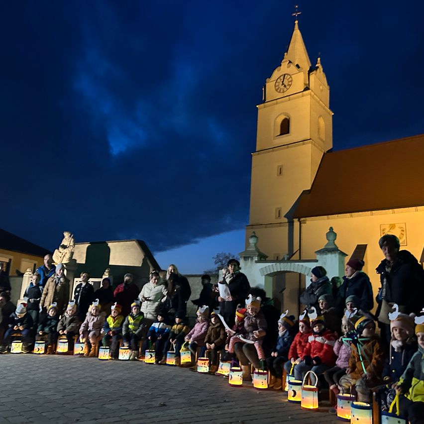 Bei Dämmerung versammeln sich eine Gruppe von Kindern und Erwachsenen vor einer Kirche, halten Laternen und tragen Mützen mit Ohren. Im Hintergrund ist ein Turm mit einer Uhr zu sehen.