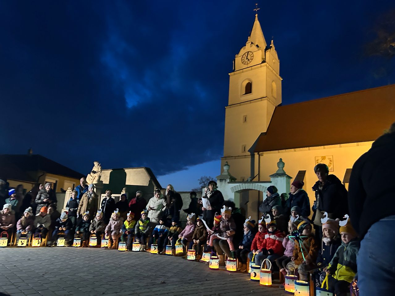 Bei Dämmerung versammeln sich eine Gruppe von Kindern und Erwachsenen vor einer Kirche, halten Laternen und tragen Mützen mit Ohren. Im Hintergrund ist ein Turm mit einer Uhr zu sehen.